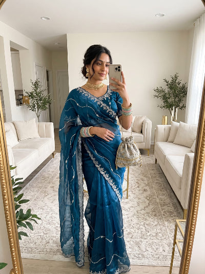 A woman takes a mirror selfie in a bright living room wearing a blue sari with silver embroidery. She is holding a silver phone and carrying a small silver pouch bag.