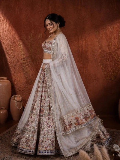 A woman models a traditional Indian lehenga choli set with a dupatta. The outfit features intricate floral embroidery in shades of brown, beige, and gold on a white base, set against a rustic terracotta wall background.