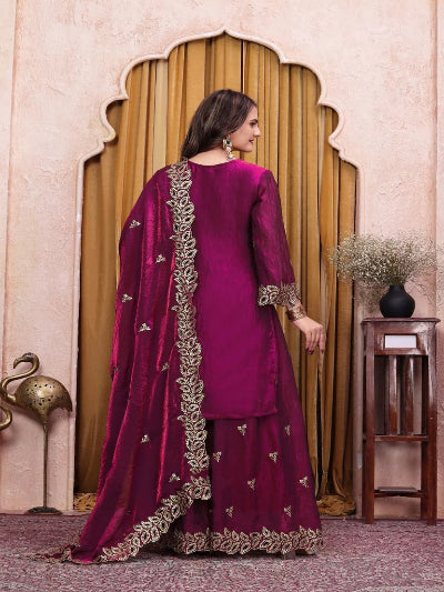 A woman wearing a traditional Indian salwar kameez suit in deep magenta with gold embroidery, shown from the back in an indoor setting with an arched wooden door.