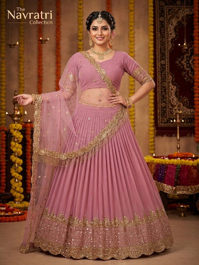 A woman models a pink and gold embroidered Indian ethnic lehenga choli set in a decorated indoor setting with marigold garlands. The set features a pink flared skirt with gold embroidery, a matching pink blouse with gold embroidery, and a sheer pink dupatta with gold borders.