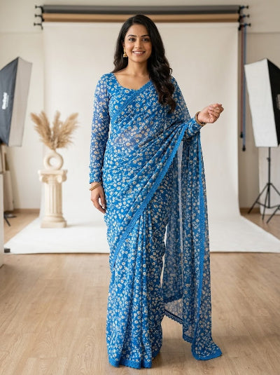 A woman modeling a blue floral print saree with a matching long-sleeve blouse in a studio setting with a white backdrop and softbox lights.