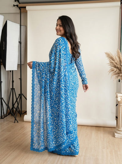 A woman modeling a bright blue long-sleeve dress with a white floral print, standing in a studio with a white backdrop and photography equipment visible on the left.