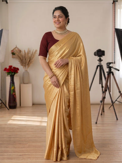 A woman modeling a golden yellow silk saree with a dark maroon short-sleeve blouse, standing in a photography studio with visible softbox lights and a tripod in the background.