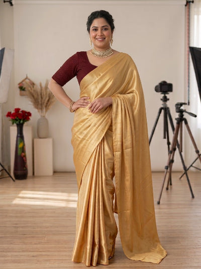 A woman modeling a golden yellow silk saree with a deep maroon short-sleeve blouse, standing in a studio with photography equipment in the background.