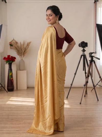 A woman modeling a golden yellow silk saree with a deep maroon short-sleeve blouse, standing in a studio with cameras and lighting equipment visible in the background.