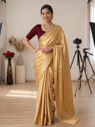 A woman modeling a golden silk saree with a deep maroon short-sleeve blouse, standing in a studio with photography equipment in the background.