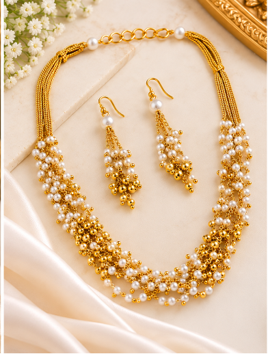 A gold and white pearl jewelry set displayed on a cream jewelry bust and draped fabric, including a multi-strand necklace, matching drop earrings, and a chain with a clasp, shown in a clean styled product layout.