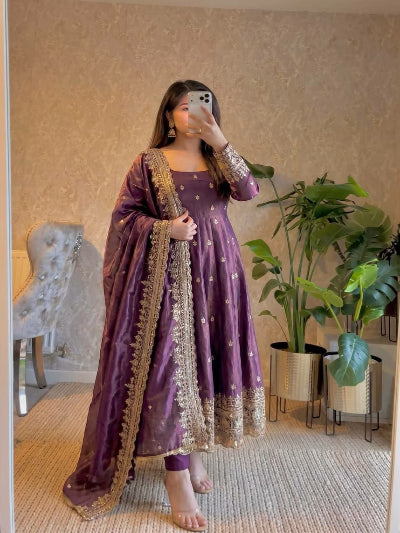 Woman in a purple traditional outfit with gold embroidery standing in a room with plants.