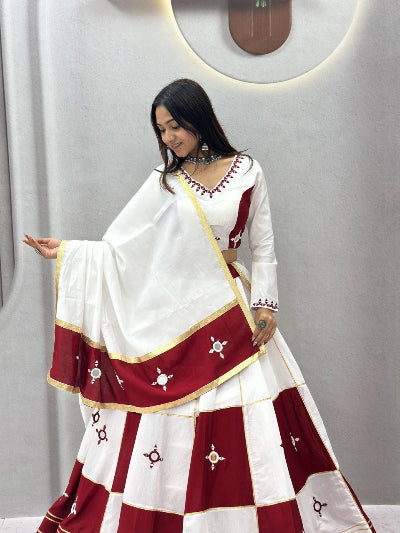 A woman modeling a traditional Indian lehenga choli set in white and deep red with gold borders and small embroidered motifs, posed against a gray wall with a circular light fixture.