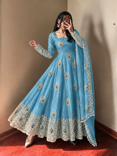 A woman wearing a light blue Anarkali gown with silver and gold mirror work embroidery and a matching dupatta, standing in an indoor setting with a beige wall and wooden floor.