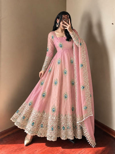 A woman wearing a pink traditional Anarkali gown with intricate gold embroidery and peacock-eye motifs, paired with a matching sheer dupatta, standing in a room corner.