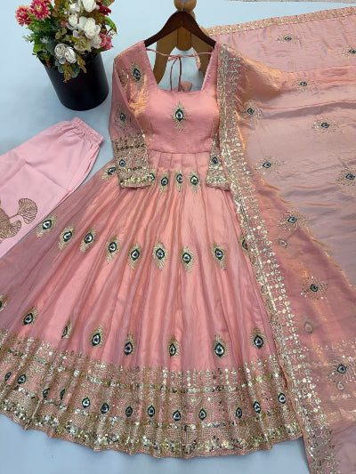 A pink traditional Anarkali gown with gold and green peacock feather embroidery, displayed with a matching dupatta and pants on a light blue background.