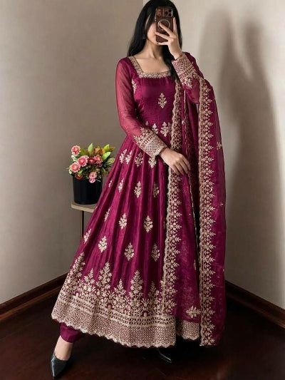 A woman wearing a deep magenta purple anarkali suit with intricate gold embroidery, taking a mirror selfie in an indoor setting with a wooden floor and beige wall.