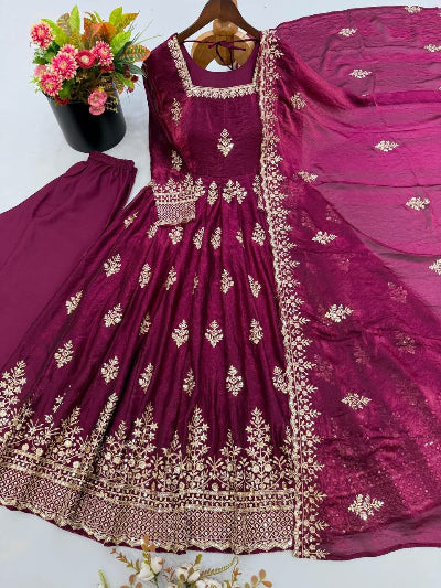 A deep magenta purple anarkali dress set with intricate gold embroidery, displayed on a hanger with matching pants and a dupatta, alongside a decorative flower pot.