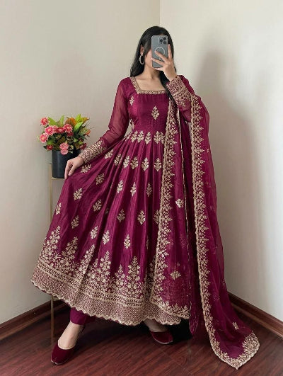 A woman takes a mirror selfie wearing a deep magenta purple Anarkali gown with intricate gold embroidery and a matching sheer dupatta draped over her shoulder, set against a plain indoor wall with a small flower arrangement.
