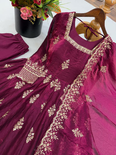 A deep magenta pink traditional dress with intricate gold embroidery, displayed on a wooden hanger with matching pants and a dupatta, set against a light background with a floral arrangement in the corner.
