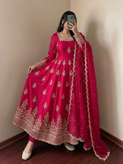 A woman takes a mirror selfie wearing a bright pink traditional Indian anarkali gown with gold embroidery and a matching dupatta, set against a plain beige wall and dark wood floor.
