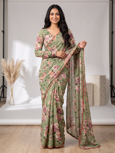 A woman modeling a sage green floral saree with a matching long-sleeve blouse in a studio setting. The saree features pink and red floral patterns with a red border, and she holds out the dupatta.