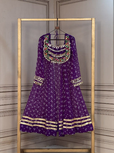 A purple Anarkali gown with white floral prints, gold zari borders, and colorful embroidered details around the neckline, hanging on a gold-framed clothing rack against a gray paneled wall.