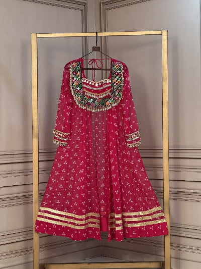 A bright pink Anarkali kurta with white floral prints, gold zari borders, and a heavily embellished neckline with green, red, and gold sequins, hanging on a gold-framed clothing rack.