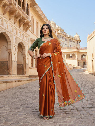 Woman in a traditional orange saree with green blouse standing in front of an architectural building.