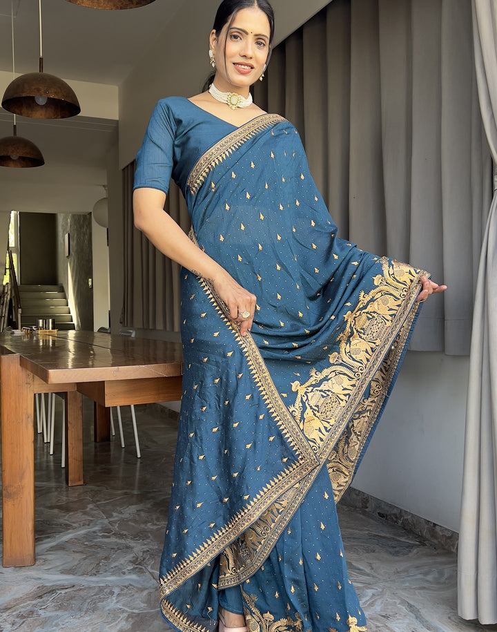 Woman in a blue saree with gold patterns standing in a room with a dining table and chairs.