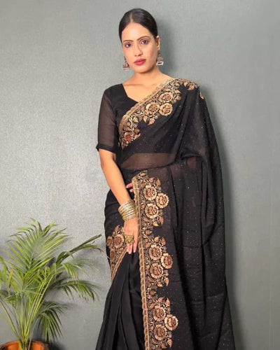 A woman models a black saree with intricate gold floral embroidery along the border and pallu, paired with a matching black sheer blouse. She is standing against a textured gray wall with a green potted plant in the lower-left corner.