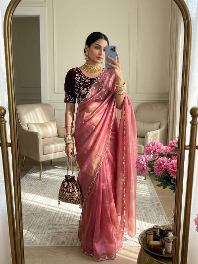 A woman takes a mirror selfie in a bright room wearing a pink sari with a dark maroon embroidered blouse, holding a phone. The sari has a sheer, semi-transparent texture with subtle gold zari detailing along the border and a pink gradient effect.