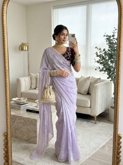A woman takes a mirror selfie in a bright living room wearing a light lavender purple saree with a dark embroidered blouse. She is holding a small cream-colored woven handbag.