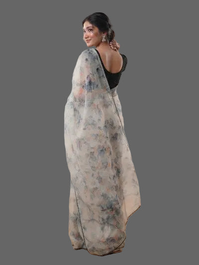 A woman wearing a semi-sheer white saree with a blue and beige floral print, paired with a black sleeveless top, standing in a studio setting with softbox lights and wooden flooring.