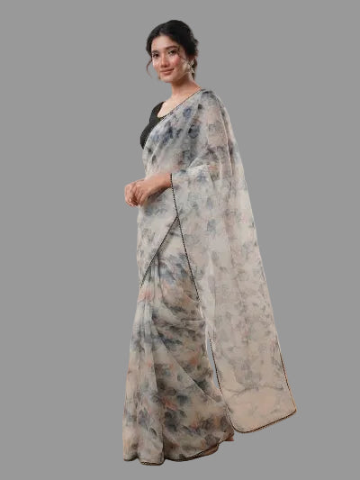 A woman models a semi-sheer white saree with a blue and gray floral print, paired with a black blouse, in a studio setting with softbox lights and wooden flooring.
