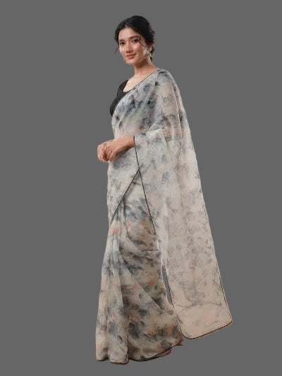 A woman modeling a semi-sheer white saree with a blue and beige floral print, paired with a black blouse, in a studio setting with softbox lights and wooden flooring.