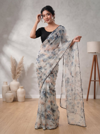 A woman models a semi-sheer floral saree with a black blouse in a styled indoor setting with neutral-colored walls, wooden floors, and decorative vases.